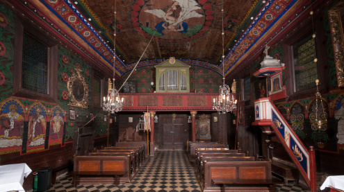 Fragment wnętrza drewnianego kościoła od strony wschodniej (prezbiterium) na chór z prospektem organowym. Całość pokryta barwną polichromią z dominującymi kolorami czerwonym, niebieskim, białym i zielonym. Po prawej stronie ambona ze schodami.