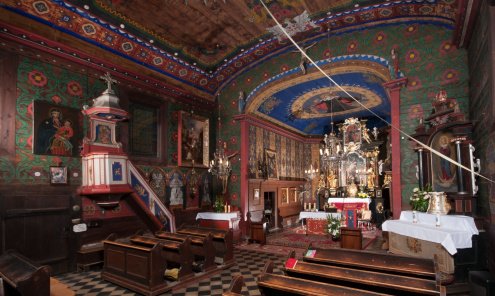 Fragment wnętrza drewnianego kościoła od strony południowo zachodniej ukazującej węższe prezbiterium i ambonę na ścianie nawy. Całość pokryta barwną polichromią z dominującym kolorem granatowym na sklepieniu prezbiterium oraz czerwonym i zielonym w nawie