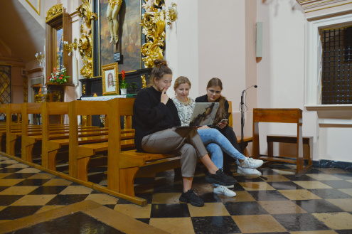 Trzy kobiety siedzą w ławce w nawie głównej kościoła, patrzą w ekran laptopa leżącego na kolanach jednej z nich.