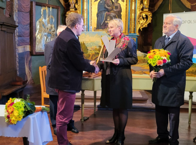Wręczenie nagrody w drewnianym kościele. Trzy osoby na tle ołtarza, dwie ściskają sobie ręce: Jóżef Skrabski i Iwona Gibas, trzecia trzyma bukiet kwiatów.