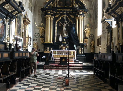 Wnętrze kościoła z widokiem na prezbiterium z ołtarzem głównym i stallami. Po lewej stronie stoi mężczyzna robiący zdjęcie aparatem fotograficznym. Na środku stoi wysoki statyw z lampą i widoczny jest pomarańczowy kabel na posadzce.