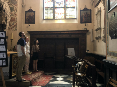 Wnętrze kaplicy bocznej kościoła z widocznym ołtarzem, konfesjonałem i witrażem. Dwie kobiety i mężczyzna stoją tyłem do ołtarza i patrzą na wysoko zawieszony obraz na przeciwległeś ścianie.