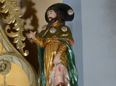Rzeźba przedstawiająca św. Rocha w stroju pielgrzyma z kapeluszem i muszlami przytwierdzonymi do pelerynki. Święty odsłania lewą nogę, na której ma ranę. Obok niego pies z bochenkiem chleba w pysku.