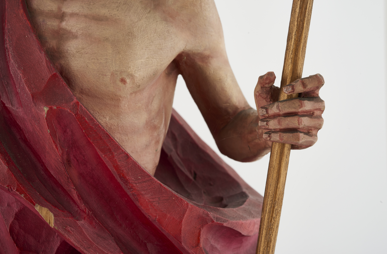 Fragment rzeźby „Chrystus Zmartwychwstały”, ukazujący klatkę piersiową Chrystusa, przewiązaną czerwonym płaszczem i lewą rękę trzymającą laskę.