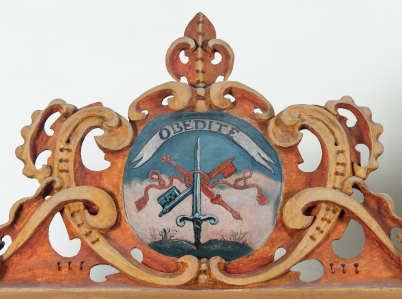 Pierwszy emblemat na szafie zakrystyjnej przedstawia miecz, skrzyżowane berło, dwa klucze oraz sznur z chwostami. Napis na szarfie głosi „OBEDITE”.