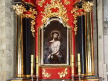 Zdjęcie ołtarza flankowanego parą kolumn. W polu głównym obraz Ecce Homo, a w zwieńczeniu obraz świętego Jana Nepomucena. Ołtarz polichromowany na kolor czerwony, detale i kolumny złocone.