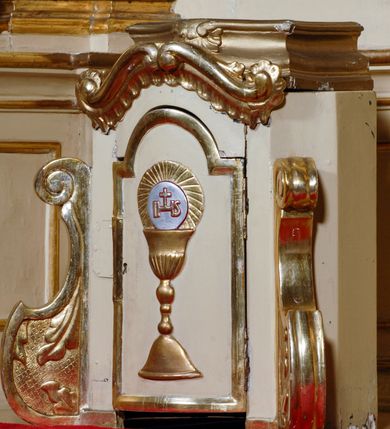 Zdjęcie nr 20: Ołtarz przyścienny, narożny, usytuowany po lewej stronie, w pierwszym od prezbiterium przęśle nawy, przy ścianie tęczowej. Architektoniczny, jednokondygnacyjny ze zwieńczeniem. Mensa murowana, prostopadłościenna, w typie skrzyniowym, z relikwiami nieznanego z imienia świętego męczennika. Antepedium z blachy w drewnianej ramie, z monogramem maryjnym w centrum. Niewielkie, drewniane tabernakulum jednodrzwiowe; lekko wypukłe drzwiczki zdobione płaskorzeźbionym kielichem z hostią wkomponowanym w złoconą płycinę zamkniętą łukiem nadwieszonym. Boki tabernakulum ujęte wolutowymi esownicami ustawionymi ukośnie w stosunku do jego lica. Zwieńczenie w formie przeciwstawnych wolut zdobionych rocaille'em. Nastawa ołtarzowa wsparta na flankujących mensę wysokich, dwudzielnych postumentach dekorowanych złoconymi płycinami zdobionymi rocaille'em. Części boczne na całej wysokości ustawione ukośnie w stosunku do osi środkowej ołtarza. Wysunięte przed zdwojone pilastry z trzonami dekorowanymi rocaille'ami, kolumny o kanelowanych na całej wysokości, złoconych trzonach i kompozytowych kapitelach, dźwigają bogato profilowane, gierowane, pełne belkowanie, wyłamane na osi podpór. Wieńczy je przerwany, półkolisty przyczółek. W polu nastawy złocona, profilowana rama o kształcie stojącego prostokąta zamkniętego łukiem wklęsło-wypukłym z uskokiem. Na jej boku, z prawej strony nałożona złocona aplikacja kwiatowa. Wewnątrz wysuwane dwa obrazy: z Matką Boską Niepokalanie Poczętą i Aniołem Stróżem (obecnie wyjęty) pędzla Wojciecha Eljasza-Radzikowskiego. Na belkowaniu osadzony rocaille'owy kartusz. Zwieńczenie o wykrojonych wolutowo bokach zamknięte łukiem wklęsło-wypukłym utworzonym przez dwie przeciwstawne woluty. U szczytu złocona, ażurowo wycinana, muszla palmetowa. W polu obraz na blasze ze św. Antonim Padewskim i Dzieciątkiem Jezus w kształcie stojącego prostokąta o ściętych krawędziach dolnych i wklęsłych bokach, zamkniętego łukiem wklęsło-wypukłym z uskokiem. Flankują go dwie, niewielkie aplikacje rocaille'owe. Na tylnych postumentach cokołu dwie dużych rozmiarów, pełnoplastyczne rzeźby aniołów. Postaci stojące w silnym kontrapoście, o tanecznych, dynamicznych pozach. Ciała muskularne, odziane w bogate, złocone, mocno fałdowane szaty. Skrzydła złocone, karnacje polichromowane. Po obu bokach ołtarza niewielkie uszy utworzone z rocaille'ów. Ołtarz obecnie przemalowany na kremowo. Ornamenty, załamania profili, ramy, trzony kolumn złocone.