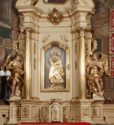 Zdjęcie nr 1: Ołtarz przyścienny, narożny, usytuowany po lewej stronie, w pierwszym od prezbiterium przęśle nawy, przy ścianie tęczowej. Architektoniczny, jednokondygnacyjny ze zwieńczeniem. Mensa murowana, prostopadłościenna, w typie skrzyniowym, z relikwiami nieznanego z imienia świętego męczennika. Antepedium z blachy w drewnianej ramie, z monogramem maryjnym w centrum. Niewielkie, drewniane tabernakulum jednodrzwiowe; lekko wypukłe drzwiczki zdobione płaskorzeźbionym kielichem z hostią wkomponowanym w złoconą płycinę zamkniętą łukiem nadwieszonym. Boki tabernakulum ujęte wolutowymi esownicami ustawionymi ukośnie w stosunku do jego lica. Zwieńczenie w formie przeciwstawnych wolut zdobionych rocaille'em. Nastawa ołtarzowa wsparta na flankujących mensę wysokich, dwudzielnych postumentach dekorowanych złoconymi płycinami zdobionymi rocaille'em. Części boczne na całej wysokości ustawione ukośnie w stosunku do osi środkowej ołtarza. Wysunięte przed zdwojone pilastry z trzonami dekorowanymi rocaille'ami, kolumny o kanelowanych na całej wysokości, złoconych trzonach i kompozytowych kapitelach, dźwigają bogato profilowane, gierowane, pełne belkowanie, wyłamane na osi podpór. Wieńczy je przerwany, półkolisty przyczółek. W polu nastawy złocona, profilowana rama o kształcie stojącego prostokąta zamkniętego łukiem wklęsło-wypukłym z uskokiem. Na jej boku, z prawej strony nałożona złocona aplikacja kwiatowa. Wewnątrz wysuwane dwa obrazy: z Matką Boską Niepokalanie Poczętą i Aniołem Stróżem (obecnie wyjęty) pędzla Wojciecha Eljasza-Radzikowskiego. Na belkowaniu osadzony rocaille'owy kartusz. Zwieńczenie o wykrojonych wolutowo bokach zamknięte łukiem wklęsło-wypukłym utworzonym przez dwie przeciwstawne woluty. U szczytu złocona, ażurowo wycinana, muszla palmetowa. W polu obraz na blasze ze św. Antonim Padewskim i Dzieciątkiem Jezus w kształcie stojącego prostokąta o ściętych krawędziach dolnych i wklęsłych bokach, zamkniętego łukiem wklęsło-wypukłym z uskokiem. Flankują go dwie, niewielkie aplikacje rocaille'owe. Na tylnych postumentach cokołu dwie dużych rozmiarów, pełnoplastyczne rzeźby aniołów. Postaci stojące w silnym kontrapoście, o tanecznych, dynamicznych pozach. Ciała muskularne, odziane w bogate, złocone, mocno fałdowane szaty. Skrzydła złocone, karnacje polichromowane. Po obu bokach ołtarza niewielkie uszy utworzone z rocaille'ów. Ołtarz obecnie przemalowany na kremowo. Ornamenty, załamania profili, ramy, trzony kolumn złocone.