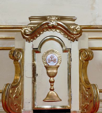 Zdjęcie nr 19: Ołtarz przyścienny, narożny, usytuowany po lewej stronie, w pierwszym od prezbiterium przęśle nawy, przy ścianie tęczowej. Architektoniczny, jednokondygnacyjny ze zwieńczeniem. Mensa murowana, prostopadłościenna, w typie skrzyniowym, z relikwiami nieznanego z imienia świętego męczennika. Antepedium z blachy w drewnianej ramie, z monogramem maryjnym w centrum. Niewielkie, drewniane tabernakulum jednodrzwiowe; lekko wypukłe drzwiczki zdobione płaskorzeźbionym kielichem z hostią wkomponowanym w złoconą płycinę zamkniętą łukiem nadwieszonym. Boki tabernakulum ujęte wolutowymi esownicami ustawionymi ukośnie w stosunku do jego lica. Zwieńczenie w formie przeciwstawnych wolut zdobionych rocaille'em. Nastawa ołtarzowa wsparta na flankujących mensę wysokich, dwudzielnych postumentach dekorowanych złoconymi płycinami zdobionymi rocaille'em. Części boczne na całej wysokości ustawione ukośnie w stosunku do osi środkowej ołtarza. Wysunięte przed zdwojone pilastry z trzonami dekorowanymi rocaille'ami, kolumny o kanelowanych na całej wysokości, złoconych trzonach i kompozytowych kapitelach, dźwigają bogato profilowane, gierowane, pełne belkowanie, wyłamane na osi podpór. Wieńczy je przerwany, półkolisty przyczółek. W polu nastawy złocona, profilowana rama o kształcie stojącego prostokąta zamkniętego łukiem wklęsło-wypukłym z uskokiem. Na jej boku, z prawej strony nałożona złocona aplikacja kwiatowa. Wewnątrz wysuwane dwa obrazy: z Matką Boską Niepokalanie Poczętą i Aniołem Stróżem (obecnie wyjęty) pędzla Wojciecha Eljasza-Radzikowskiego. Na belkowaniu osadzony rocaille'owy kartusz. Zwieńczenie o wykrojonych wolutowo bokach zamknięte łukiem wklęsło-wypukłym utworzonym przez dwie przeciwstawne woluty. U szczytu złocona, ażurowo wycinana, muszla palmetowa. W polu obraz na blasze ze św. Antonim Padewskim i Dzieciątkiem Jezus w kształcie stojącego prostokąta o ściętych krawędziach dolnych i wklęsłych bokach, zamkniętego łukiem wklęsło-wypukłym z uskokiem. Flankują go dwie, niewielkie aplikacje rocaille'owe. Na tylnych postumentach cokołu dwie dużych rozmiarów, pełnoplastyczne rzeźby aniołów. Postaci stojące w silnym kontrapoście, o tanecznych, dynamicznych pozach. Ciała muskularne, odziane w bogate, złocone, mocno fałdowane szaty. Skrzydła złocone, karnacje polichromowane. Po obu bokach ołtarza niewielkie uszy utworzone z rocaille'ów. Ołtarz obecnie przemalowany na kremowo. Ornamenty, załamania profili, ramy, trzony kolumn złocone.
