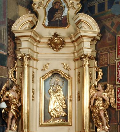 Zdjęcie nr 2: Ołtarz przyścienny, narożny, usytuowany po lewej stronie, w pierwszym od prezbiterium przęśle nawy, przy ścianie tęczowej. Architektoniczny, jednokondygnacyjny ze zwieńczeniem. Mensa murowana, prostopadłościenna, w typie skrzyniowym, z relikwiami nieznanego z imienia świętego męczennika. Antepedium z blachy w drewnianej ramie, z monogramem maryjnym w centrum. Niewielkie, drewniane tabernakulum jednodrzwiowe; lekko wypukłe drzwiczki zdobione płaskorzeźbionym kielichem z hostią wkomponowanym w złoconą płycinę zamkniętą łukiem nadwieszonym. Boki tabernakulum ujęte wolutowymi esownicami ustawionymi ukośnie w stosunku do jego lica. Zwieńczenie w formie przeciwstawnych wolut zdobionych rocaille'em. Nastawa ołtarzowa wsparta na flankujących mensę wysokich, dwudzielnych postumentach dekorowanych złoconymi płycinami zdobionymi rocaille'em. Części boczne na całej wysokości ustawione ukośnie w stosunku do osi środkowej ołtarza. Wysunięte przed zdwojone pilastry z trzonami dekorowanymi rocaille'ami, kolumny o kanelowanych na całej wysokości, złoconych trzonach i kompozytowych kapitelach, dźwigają bogato profilowane, gierowane, pełne belkowanie, wyłamane na osi podpór. Wieńczy je przerwany, półkolisty przyczółek. W polu nastawy złocona, profilowana rama o kształcie stojącego prostokąta zamkniętego łukiem wklęsło-wypukłym z uskokiem. Na jej boku, z prawej strony nałożona złocona aplikacja kwiatowa. Wewnątrz wysuwane dwa obrazy: z Matką Boską Niepokalanie Poczętą i Aniołem Stróżem (obecnie wyjęty) pędzla Wojciecha Eljasza-Radzikowskiego. Na belkowaniu osadzony rocaille'owy kartusz. Zwieńczenie o wykrojonych wolutowo bokach zamknięte łukiem wklęsło-wypukłym utworzonym przez dwie przeciwstawne woluty. U szczytu złocona, ażurowo wycinana, muszla palmetowa. W polu obraz na blasze ze św. Antonim Padewskim i Dzieciątkiem Jezus w kształcie stojącego prostokąta o ściętych krawędziach dolnych i wklęsłych bokach, zamkniętego łukiem wklęsło-wypukłym z uskokiem. Flankują go dwie, niewielkie aplikacje rocaille'owe. Na tylnych postumentach cokołu dwie dużych rozmiarów, pełnoplastyczne rzeźby aniołów. Postaci stojące w silnym kontrapoście, o tanecznych, dynamicznych pozach. Ciała muskularne, odziane w bogate, złocone, mocno fałdowane szaty. Skrzydła złocone, karnacje polichromowane. Po obu bokach ołtarza niewielkie uszy utworzone z rocaille'ów. Ołtarz obecnie przemalowany na kremowo. Ornamenty, załamania profili, ramy, trzony kolumn złocone.