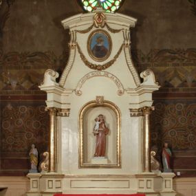 Zdjęcie nr 1: Ołtarz, architektoniczny, przyścienny, jednoosiowy, jednokondygnacyjny ze zwieńczeniem.  Mensa drewniana w postaci stołu wspartego na czterech nogach naśladujących w formie filary z impostowymi kapitelami. Retabulum posadowione na wysokim, dwudzielnym, rozczłonkowanym architektonicznie cokole, dekorowanym prostokątnymi płycinami o złoconych brzegach. Przednie ramiona cokołu ustawione ukośnie w stosunku do osi ołtarza. Na nich kolumny o kompozytowych kapitelach, wysunięte przed pilastry z pseudokorynckimi kapitelami i kanelurowanymi trzonami. Ujęte dołem parą wolut podpory dźwigają wyłamane ponad nimi, pełne belkowanie, przerwane na osi ołtarza. Na nim odcinki przyczółka w postaci wolut. Na architraw belkowania, ponad kapitelami kolumn nałożone pojedyncze liście akantu. Pomiędzy partiami przerwanego belkowania banderola rozpięta w formie półkolistego łuku, z napisem „ŚW(IĘTY) ANTONI MÓDL SIĘ ZA NAMI!". Pola odcinków przyczółka dekorowane złoconymi rocaille'ami. W polu ołtarza prostokątna nisza zamknięta łukiem koszowym z wydatnym kluczem w zwieńczeniu, ujęta złoconą, profilowaną ramą dekorowaną perełkowaniem. W niej pełnoplastyczna figura św. Antoniego Padewskiego. Wysokie, dużych rozmiarów trapezoidalne, ujęte po bokach motywem taśm zwieńczenie zamknięte fragmentem gzymsu w kształcie łuku odcinkowego, na osi którego kartusz wolutowy z motywem gorejącego serca pośrodku. Boki zwieńczenia w górnej partii ujęte parą wolut. Szczyt w formie krzyża na cokole z pary wolutek. W polu owalny obraz w złoconej, profilowanej ramie przedstawiający niezidentyfikowaną świętą. Wizerunek ujmują festony. Po bokach nastawy para figur św. Piotra i św. Pawła na wysuniętych partiach cokołu nastawy. Ołtarz w kolorze białym. Złocenia w partiach ornamentu oraz detalu architektonicznego.