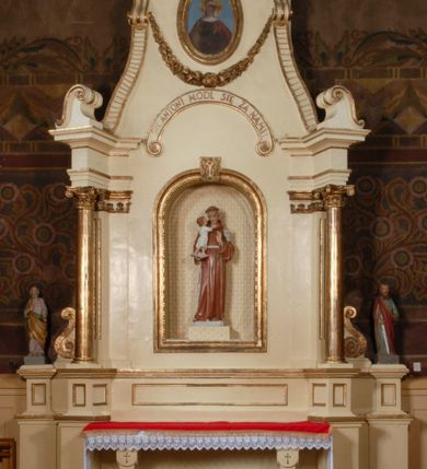 Zdjęcie nr 2: Ołtarz, architektoniczny, przyścienny, jednoosiowy, jednokondygnacyjny ze zwieńczeniem.  Mensa drewniana w postaci stołu wspartego na czterech nogach naśladujących w formie filary z impostowymi kapitelami. Retabulum posadowione na wysokim, dwudzielnym, rozczłonkowanym architektonicznie cokole, dekorowanym prostokątnymi płycinami o złoconych brzegach. Przednie ramiona cokołu ustawione ukośnie w stosunku do osi ołtarza. Na nich kolumny o kompozytowych kapitelach, wysunięte przed pilastry z pseudokorynckimi kapitelami i kanelurowanymi trzonami. Ujęte dołem parą wolut podpory dźwigają wyłamane ponad nimi, pełne belkowanie, przerwane na osi ołtarza. Na nim odcinki przyczółka w postaci wolut. Na architraw belkowania, ponad kapitelami kolumn nałożone pojedyncze liście akantu. Pomiędzy partiami przerwanego belkowania banderola rozpięta w formie półkolistego łuku, z napisem „ŚW(IĘTY) ANTONI MÓDL SIĘ ZA NAMI!". Pola odcinków przyczółka dekorowane złoconymi rocaille'ami. W polu ołtarza prostokątna nisza zamknięta łukiem koszowym z wydatnym kluczem w zwieńczeniu, ujęta złoconą, profilowaną ramą dekorowaną perełkowaniem. W niej pełnoplastyczna figura św. Antoniego Padewskiego. Wysokie, dużych rozmiarów trapezoidalne, ujęte po bokach motywem taśm zwieńczenie zamknięte fragmentem gzymsu w kształcie łuku odcinkowego, na osi którego kartusz wolutowy z motywem gorejącego serca pośrodku. Boki zwieńczenia w górnej partii ujęte parą wolut. Szczyt w formie krzyża na cokole z pary wolutek. W polu owalny obraz w złoconej, profilowanej ramie przedstawiający niezidentyfikowaną świętą. Wizerunek ujmują festony. Po bokach nastawy para figur św. Piotra i św. Pawła na wysuniętych partiach cokołu nastawy. Ołtarz w kolorze białym. Złocenia w partiach ornamentu oraz detalu architektonicznego.