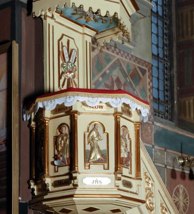 Zdjęcie nr 1: Ambona umieszczona na północnej ścianie, drugiego od prezbiterium przęsła nawy, przy lewym ramieniu transeptu. Ambona formie pięciobocznego kosza ze stożkowatą podstawą, zapleckiem i baldachimem zwieńczonym pełnoplastyczną rzeźbą archanioła dmącego w trąbę. Dostępna od wschodu – prowadzą do niej wysokie, trójdzielne schody z drewnianą balustradą. Dodatkowe drzwi znajdują się w ścianie północnej nawy, na prawo i nieco poniżej zaplecka. Stożkowata podstawa z polami dekorowanymi płaskorzeźbionymi, złoconymi rombami oddzielona od kosza za pomocą bogato profilowanego gzymsu. Kosz artykułowany za pomocą złoconych kolumienek na wolutowych postumentach, dźwigających odcinki niepełnego belkowania. W polach prostokątne płyciny o podciętych schodkowo podstawach zwieńczone łukiem nadwieszonym. W nich płaskorzeźbione przedstawienia od lewej: św. Jana z orłem piszącego piórem w księdze, stojącego na tle Golgoty, poniżej prostokątna tablica o ściętych narożach z napisem „Św(ięty) JAN”; św. Łukasza z piórem, kałamarzem i zamkniętą księgą w rękach, u jego stóp uskrzydlona głowa wołu, poniżej tablica z napisem „Św. ŁUKASZ”; Chrystusa siewcy ukazanego na tle pejzażu, poniżej tablica z monogramem „IHS”; św. Marka z księgą i piórem, uskrzydlony lew u jego stóp, poniżej tablica z napisem: „Św. MAREK”; św. Mateusza z piórem i księgą w ręku i klęczącym aniołem u jego stóp, poniżej tablica z napisem „Św(ięty) MATEUSZ”. Powyżej, w partiach fryzu analogiczne tablice z napisem „KTO JEST / Z BOGA / SŁÓW / BOŻYCH / SŁUCHA”. Kosz zwieńczony profilowanym gzymsem. Zaplecek ujęty pilastrami o kanelurowanych trzonach, w jego polu płycina w kształcie stojącego prostokąta o ściętych łukowato, wklęsłych narożach. W jej centrum płaskorzeźbione skrzyżowane tablice Dziesięciu Przykazań na srebrzonych obłokach, otoczone złoconymi promieniami. Baldachim czworoboczny zamknięty profilowanym gzymsem, pod którym podwieszona kotara z lambrekinem. Podniebie malowane na błękitny kolor z nałożoną w centrum, płaskorzeźbioną gołębicą Ducha Świętego w glorii. W zwieńczeniu ustawiona na wolutowym, rozczłonkowanym postumencie figura archanioła z trąbą. Rzeźba pełnoplastyczna, złocona, polichromowana. Postać wygięta łukowato, w prawej ręce wzniesionej ku górze trzyma długą trąbę. Lewej ręki brak. Anioł ubrany w długie, rozwiane, miękko układające się szaty przepasane sznurem. Twarz podłużna z wydatnymi policzkami, małym wystającym podbródkiem i dużymi, niebieskimi oczyma. Włosy sięgające ramion, kręcone, koloru brązowego.
Schody drewniane, trójdzielne z drewnianą balustradą zdobioną płycinami, w których nałożone, złocone aplikacje z silnie przestylizowanego akantu. Poręcz złocona zakończona niewielką wolutą.
Ambona malowana na kremowo, ze złoceniami w partiach ornamentów, załomach elementów architektonicznych oraz na szatach postaci.
