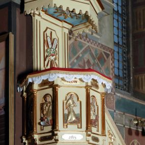 Zdjęcie nr 1: Ambona umieszczona na północnej ścianie, drugiego od prezbiterium przęsła nawy, przy lewym ramieniu transeptu. Ambona formie pięciobocznego kosza ze stożkowatą podstawą, zapleckiem i baldachimem zwieńczonym pełnoplastyczną rzeźbą archanioła dmącego w trąbę. Dostępna od wschodu – prowadzą do niej wysokie, trójdzielne schody z drewnianą balustradą. Dodatkowe drzwi znajdują się w ścianie północnej nawy, na prawo i nieco poniżej zaplecka. Stożkowata podstawa z polami dekorowanymi płaskorzeźbionymi, złoconymi rombami oddzielona od kosza za pomocą bogato profilowanego gzymsu. Kosz artykułowany za pomocą złoconych kolumienek na wolutowych postumentach, dźwigających odcinki niepełnego belkowania. W polach prostokątne płyciny o podciętych schodkowo podstawach zwieńczone łukiem nadwieszonym. W nich płaskorzeźbione przedstawienia od lewej: św. Jana z orłem piszącego piórem w księdze, stojącego na tle Golgoty, poniżej prostokątna tablica o ściętych narożach z napisem „Św(ięty) JAN”; św. Łukasza z piórem, kałamarzem i zamkniętą księgą w rękach, u jego stóp uskrzydlona głowa wołu, poniżej tablica z napisem „Św. ŁUKASZ”; Chrystusa siewcy ukazanego na tle pejzażu, poniżej tablica z monogramem „IHS”; św. Marka z księgą i piórem, uskrzydlony lew u jego stóp, poniżej tablica z napisem: „Św. MAREK”; św. Mateusza z piórem i księgą w ręku i klęczącym aniołem u jego stóp, poniżej tablica z napisem „Św(ięty) MATEUSZ”. Powyżej, w partiach fryzu analogiczne tablice z napisem „KTO JEST / Z BOGA / SŁÓW / BOŻYCH / SŁUCHA”. Kosz zwieńczony profilowanym gzymsem. Zaplecek ujęty pilastrami o kanelurowanych trzonach, w jego polu płycina w kształcie stojącego prostokąta o ściętych łukowato, wklęsłych narożach. W jej centrum płaskorzeźbione skrzyżowane tablice Dziesięciu Przykazań na srebrzonych obłokach, otoczone złoconymi promieniami. Baldachim czworoboczny zamknięty profilowanym gzymsem, pod którym podwieszona kotara z lambrekinem. Podniebie malowane na błękitny kolor z nałożoną w centrum, płaskorzeźbioną gołębicą Ducha Świętego w glorii. W zwieńczeniu ustawiona na wolutowym, rozczłonkowanym postumencie figura archanioła z trąbą. Rzeźba pełnoplastyczna, złocona, polichromowana. Postać wygięta łukowato, w prawej ręce wzniesionej ku górze trzyma długą trąbę. Lewej ręki brak. Anioł ubrany w długie, rozwiane, miękko układające się szaty przepasane sznurem. Twarz podłużna z wydatnymi policzkami, małym wystającym podbródkiem i dużymi, niebieskimi oczyma. Włosy sięgające ramion, kręcone, koloru brązowego.
Schody drewniane, trójdzielne z drewnianą balustradą zdobioną płycinami, w których nałożone, złocone aplikacje z silnie przestylizowanego akantu. Poręcz złocona zakończona niewielką wolutą.
Ambona malowana na kremowo, ze złoceniami w partiach ornamentów, załomach elementów architektonicznych oraz na szatach postaci.