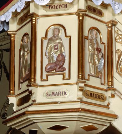 Zdjęcie nr 8: Ambona umieszczona na północnej ścianie, drugiego od prezbiterium przęsła nawy, przy lewym ramieniu transeptu. Ambona formie pięciobocznego kosza ze stożkowatą podstawą, zapleckiem i baldachimem zwieńczonym pełnoplastyczną rzeźbą archanioła dmącego w trąbę. Dostępna od wschodu – prowadzą do niej wysokie, trójdzielne schody z drewnianą balustradą. Dodatkowe drzwi znajdują się w ścianie północnej nawy, na prawo i nieco poniżej zaplecka. Stożkowata podstawa z polami dekorowanymi płaskorzeźbionymi, złoconymi rombami oddzielona od kosza za pomocą bogato profilowanego gzymsu. Kosz artykułowany za pomocą złoconych kolumienek na wolutowych postumentach, dźwigających odcinki niepełnego belkowania. W polach prostokątne płyciny o podciętych schodkowo podstawach zwieńczone łukiem nadwieszonym. W nich płaskorzeźbione przedstawienia od lewej: św. Jana z orłem piszącego piórem w księdze, stojącego na tle Golgoty, poniżej prostokątna tablica o ściętych narożach z napisem „Św(ięty) JAN”; św. Łukasza z piórem, kałamarzem i zamkniętą księgą w rękach, u jego stóp uskrzydlona głowa wołu, poniżej tablica z napisem „Św. ŁUKASZ”; Chrystusa siewcy ukazanego na tle pejzażu, poniżej tablica z monogramem „IHS”; św. Marka z księgą i piórem, uskrzydlony lew u jego stóp, poniżej tablica z napisem: „Św. MAREK”; św. Mateusza z piórem i księgą w ręku i klęczącym aniołem u jego stóp, poniżej tablica z napisem „Św(ięty) MATEUSZ”. Powyżej, w partiach fryzu analogiczne tablice z napisem „KTO JEST / Z BOGA / SŁÓW / BOŻYCH / SŁUCHA”. Kosz zwieńczony profilowanym gzymsem. Zaplecek ujęty pilastrami o kanelurowanych trzonach, w jego polu płycina w kształcie stojącego prostokąta o ściętych łukowato, wklęsłych narożach. W jej centrum płaskorzeźbione skrzyżowane tablice Dziesięciu Przykazań na srebrzonych obłokach, otoczone złoconymi promieniami. Baldachim czworoboczny zamknięty profilowanym gzymsem, pod którym podwieszona kotara z lambrekinem. Podniebie malowane na błękitny kolor z nałożoną w centrum, płaskorzeźbioną gołębicą Ducha Świętego w glorii. W zwieńczeniu ustawiona na wolutowym, rozczłonkowanym postumencie figura archanioła z trąbą. Rzeźba pełnoplastyczna, złocona, polichromowana. Postać wygięta łukowato, w prawej ręce wzniesionej ku górze trzyma długą trąbę. Lewej ręki brak. Anioł ubrany w długie, rozwiane, miękko układające się szaty przepasane sznurem. Twarz podłużna z wydatnymi policzkami, małym wystającym podbródkiem i dużymi, niebieskimi oczyma. Włosy sięgające ramion, kręcone, koloru brązowego.
Schody drewniane, trójdzielne z drewnianą balustradą zdobioną płycinami, w których nałożone, złocone aplikacje z silnie przestylizowanego akantu. Poręcz złocona zakończona niewielką wolutą.
Ambona malowana na kremowo, ze złoceniami w partiach ornamentów, załomach elementów architektonicznych oraz na szatach postaci.