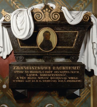Zdjęcie nr 1: Rozbudowane epitafium utworzone z wysokiego cokołu z inskrypcją i wspartego na nim sarkofagu nakrytego tkaniną z portretem. Sarkofag stoi na trzech niewielkich nóżkach, zdobionych podwójnymi ustawionymi przeciwstawnie wolutami. Ma kształt równoramiennego trapezu, z dekorowaną perełkowaniem płyciną pośrodku i osadzonym w jej centrum, malowanym na owalnej, złoconej blasze portretem zmarłego. Zwieńczenie w kształcie łuku wklęsło-wypukłego, dekorowane złoconymi wolutami zakończonymi liśćmi akantu liściastymi wolutami w funkcji akroterionów oraz narzuconą nań, udrapowaną, białą tkaniną. Pośrodku owalny obraz w złoconej ramie naśladującej węża pożerającego własny ogon. Zmarły przedstawiony w popiersiu, z ciałem w trzech czwartych zwróconym w lewo. Ubrany w ciemny strój z wysokim kołnierzem. Tło jednolicie złocone. Na cokole, wspartym na dwóch piramidalnych konsolach ryta i złocona inskrypcja "FRANCISZKOWI ŁĄCKIEMU / KTÓRY DO ZASŁUG, I CNÓT OBYWATELSKICH, / ŁĄCZYŁ DOBROCZYNNOŚĆ. / A TEY BŁOGI WPŁYW DOSIĘGNĄŁ WIELU. / W WIEKU LAT 54 D(NIA) 15 WRZEŚNIA 1844. R(OKU) ZMARŁEMU."