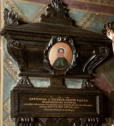 Zdjęcie nr 1: Rozbudowane epitafium utworzone z tablicy inskrypcyjnej i posadowionego na niej sarkofagu z owalnym portretem zmarłej. Tablica osadzona na dwóch wolutowych, profilowanych konsolach i umieszczonych między nimi płaskich wolutach połączonych muszlą, z inskrypcją: "D(EO) O(PTIMO) M(AXIMO) / ANTONINA Z SZYBALSKICH ŁĄCKA / WŁAŚCICIELKA LIPOWCA / zacna obywatelka wzorowa żona, najczulsza matka opiekunka ubogich, wylana dla przyjaciół / zmarła dnia 10/6 1888 w 90 roku życia. / Prosi o westchnienie do Boga." Sarkofag oparty na dwóch lwich łapach i postumencie pośrodku, wybrzuszony, ujęty liśćmi akantu, zwieńczony spłaszczoną trójkątną pokrywą z liściastymi akroterionami na bokach i trójkątnym cokolikiem z dwiema wolutkami z palmetką, zwieńczoną krzyżem. Na sarkofagu owalny medalion otoczony kartuszem z liści z portretem zmarłej. Kobieta przedstawiona jest frontalnie, w popiersiu. Ubrana w ciemno-zieloną pelerynę oraz biały czepiec zawiązywany zieloną kokardą. Tło jednolicie złocone. 