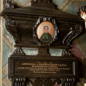Zdjęcie nr 1: Rozbudowane epitafium utworzone z tablicy inskrypcyjnej i posadowionego na niej sarkofagu z owalnym portretem zmarłej. Tablica osadzona na dwóch wolutowych, profilowanych konsolach i umieszczonych między nimi płaskich wolutach połączonych muszlą, z inskrypcją: "D(EO) O(PTIMO) M(AXIMO) / ANTONINA Z SZYBALSKICH ŁĄCKA / WŁAŚCICIELKA LIPOWCA / zacna obywatelka wzorowa żona, najczulsza matka opiekunka ubogich, wylana dla przyjaciół / zmarła dnia 10/6 1888 w 90 roku życia. / Prosi o westchnienie do Boga." Sarkofag oparty na dwóch lwich łapach i postumencie pośrodku, wybrzuszony, ujęty liśćmi akantu, zwieńczony spłaszczoną trójkątną pokrywą z liściastymi akroterionami na bokach i trójkątnym cokolikiem z dwiema wolutkami z palmetką, zwieńczoną krzyżem. Na sarkofagu owalny medalion otoczony kartuszem z liści z portretem zmarłej. Kobieta przedstawiona jest frontalnie, w popiersiu. Ubrana w ciemno-zieloną pelerynę oraz biały czepiec zawiązywany zieloną kokardą. Tło jednolicie złocone. 