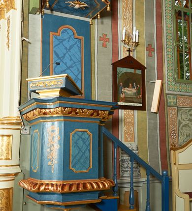 Zdjęcie nr 2: Ambona wisząca, przyścienna. Kosz na planie kwadratu o wklęsłych narożach; na profilowanej podstawie, cokół wydatny zdobiony wklęsłym puklowaniem; u spodu szyszka. Ścianki kosza dekorowane malowanymi płycinami, w formie stojących prostokątów z wklęsłymi narożami, w polu środkowym marmoryzowane zdobienia. Ścianki wklęsłe zdobione rocaille'm. Parapet w formie profilowanego, pełnego belkowania z fryzem dekorowanym motywem roślinnym. Na parapecie, w lewym narożu pulpit; od frontu zdobiony płyciną z rocaille'm. Zaplecek w formie wydłużonego, stojącego prostokąta, w polu z malowaną płyciną w kształcie stojącego prostokąta zamkniętego łukiem nadwieszonym. Płycina marmoryzowana i obwiedziona złotą ramką. Baldachim na planie kwadratu z wklęsłymi narożami; ujęty lambrekinem, zamknięty gzymsem. U spodu rzeźbiona gołębica Ducha Świętego w glorii. Wejście na ambonę od strony prezbiterium, w postaci pięciostopniowych schodów z balustradą. Struktura polichromowana na niebiesko, profile i ornamenty złocone.