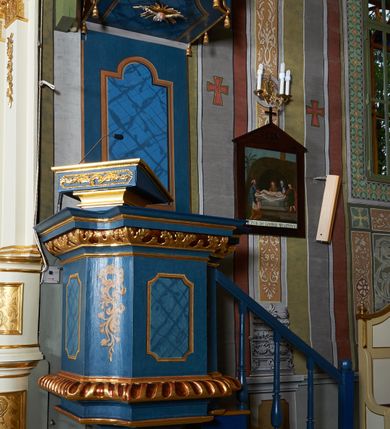 Zdjęcie nr 1: Ambona wisząca, przyścienna. Kosz na planie kwadratu o wklęsłych narożach; na profilowanej podstawie, cokół wydatny zdobiony wklęsłym puklowaniem; u spodu szyszka. Ścianki kosza dekorowane malowanymi płycinami, w formie stojących prostokątów z wklęsłymi narożami, w polu środkowym marmoryzowane zdobienia. Ścianki wklęsłe zdobione rocaille&#039;m. Parapet w formie profilowanego, pełnego belkowania z fryzem dekorowanym motywem roślinnym. Na parapecie, w lewym narożu pulpit; od frontu zdobiony płyciną z rocaille&#039;m. Zaplecek w formie wydłużonego, stojącego prostokąta, w polu z malowaną płyciną w kształcie stojącego prostokąta zamkniętego łukiem nadwieszonym. Płycina marmoryzowana i obwiedziona złotą ramką. Baldachim na planie kwadratu z wklęsłymi narożami; ujęty lambrekinem, zamknięty gzymsem. U spodu rzeźbiona gołębica Ducha Świętego w glorii. Wejście na ambonę od strony prezbiterium, w postaci pięciostopniowych schodów z balustradą. Struktura polichromowana na niebiesko, profile i ornamenty złocone.