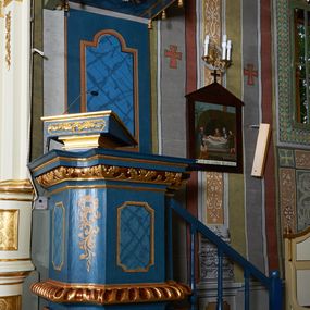 Zdjęcie nr 1: Ambona wisząca, przyścienna. Kosz na planie kwadratu o wklęsłych narożach; na profilowanej podstawie, cokół wydatny zdobiony wklęsłym puklowaniem; u spodu szyszka. Ścianki kosza dekorowane malowanymi płycinami, w formie stojących prostokątów z wklęsłymi narożami, w polu środkowym marmoryzowane zdobienia. Ścianki wklęsłe zdobione rocaille'm. Parapet w formie profilowanego, pełnego belkowania z fryzem dekorowanym motywem roślinnym. Na parapecie, w lewym narożu pulpit; od frontu zdobiony płyciną z rocaille'm. Zaplecek w formie wydłużonego, stojącego prostokąta, w polu z malowaną płyciną w kształcie stojącego prostokąta zamkniętego łukiem nadwieszonym. Płycina marmoryzowana i obwiedziona złotą ramką. Baldachim na planie kwadratu z wklęsłymi narożami; ujęty lambrekinem, zamknięty gzymsem. U spodu rzeźbiona gołębica Ducha Świętego w glorii. Wejście na ambonę od strony prezbiterium, w postaci pięciostopniowych schodów z balustradą. Struktura polichromowana na niebiesko, profile i ornamenty złocone.