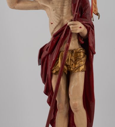 Zdjęcie nr 8: Rzeźba pełna, ustawiona na niskim, kwadratowym, profilowanym cokole. Chrystus ukazany frontalnie, stojący na kawałku zielonej murawy, w kontrapoście, z głową zwróconą w prawo. W lewej, ugiętej ręce trzyma chorągiew; prawa, uniesiona jest wysoko w geście błogosławieństwa. Twarz pociągła, okolona brodą, z na wpół przymkniętymi oczami; włosy długie, kręcone opadają na ramiona. Ciało wychudzone, z zaznaczonymi śladami męki i podkreśloną linią żeber. Przez lewe ramię przerzucony czerwony płaszcz spływający na plecy i z przodu na lewe przedramię; na biodrach złocone  perizonium, ciasno oplecione. Polichromia ciała jest naturalistyczna, perizonium złocone.