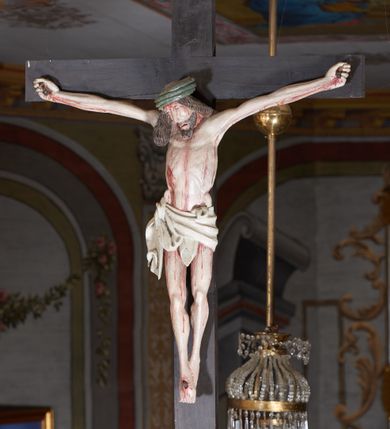Zdjęcie nr 3: Rzeźba ścięta z tyłu. Figura Chrystusa Ukrzyżowanego w lekkim zwisie, o szeroko rozłożonych ramionach. Głowa przechylona na prawe ramię. Twarz pociągła, szczupła, okolona zarostem, o niewielkich, półprzymkniętych oczach, prostym i długim nosie i otwartych ustach. Włosy w formie grubych pukli, opadających na prawe ramię. Korona cierniowa w kształcie czepca. Sylwetka ciała nienaturalnie wydłużona, szczupła, z zaznaczonymi śladami męki. Perizonium ściśle opinające biodra, obficie drapowane, zawiązane na prawym boku. Polichromia naturalistyczna. Krzyż gładki, malowany na czarno.