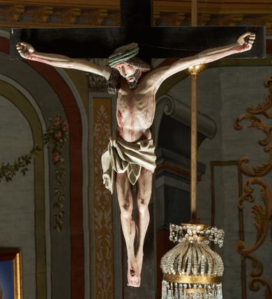 Zdjęcie nr 2: Rzeźba ścięta z tyłu. Figura Chrystusa Ukrzyżowanego w lekkim zwisie, o szeroko rozłożonych ramionach. Głowa przechylona na prawe ramię. Twarz pociągła, szczupła, okolona zarostem, o niewielkich, półprzymkniętych oczach, prostym i długim nosie i otwartych ustach. Włosy w formie grubych pukli, opadających na prawe ramię. Korona cierniowa w kształcie czepca. Sylwetka ciała nienaturalnie wydłużona, szczupła, z zaznaczonymi śladami męki. Perizonium ściśle opinające biodra, obficie drapowane, zawiązane na prawym boku. Polichromia naturalistyczna. Krzyż gładki, malowany na czarno.