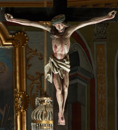 Zdjęcie nr 1: Rzeźba ścięta z tyłu. Figura Chrystusa Ukrzyżowanego w lekkim zwisie, o szeroko rozłożonych ramionach. Głowa przechylona na prawe ramię. Twarz pociągła, szczupła, okolona zarostem, o niewielkich, półprzymkniętych oczach, prostym i długim nosie i otwartych ustach. Włosy w formie grubych pukli, opadających na prawe ramię. Korona cierniowa w kształcie czepca. Sylwetka ciała nienaturalnie wydłużona, szczupła, z zaznaczonymi śladami męki. Perizonium ściśle opinające biodra, obficie drapowane, zawiązane na prawym boku. Polichromia naturalistyczna. Krzyż gładki, malowany na czarno.