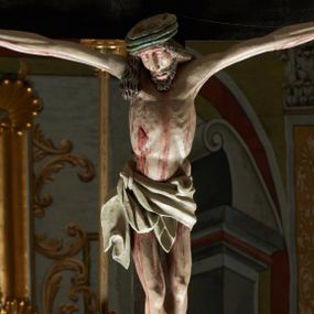 Zdjęcie nr 1: Rzeźba ścięta z tyłu. Figura Chrystusa Ukrzyżowanego w lekkim zwisie, o szeroko rozłożonych ramionach. Głowa przechylona na prawe ramię. Twarz pociągła, szczupła, okolona zarostem, o niewielkich, półprzymkniętych oczach, prostym i długim nosie i otwartych ustach. Włosy w formie grubych pukli, opadających na prawe ramię. Korona cierniowa w kształcie czepca. Sylwetka ciała nienaturalnie wydłużona, szczupła, z zaznaczonymi śladami męki. Perizonium ściśle opinające biodra, obficie drapowane, zawiązane na prawym boku. Polichromia naturalistyczna. Krzyż gładki, malowany na czarno.