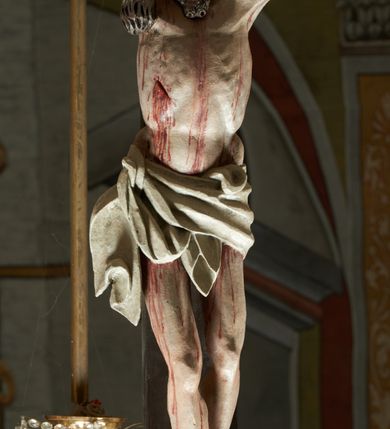 Zdjęcie nr 4: Rzeźba ścięta z tyłu. Figura Chrystusa Ukrzyżowanego w lekkim zwisie, o szeroko rozłożonych ramionach. Głowa przechylona na prawe ramię. Twarz pociągła, szczupła, okolona zarostem, o niewielkich, półprzymkniętych oczach, prostym i długim nosie i otwartych ustach. Włosy w formie grubych pukli, opadających na prawe ramię. Korona cierniowa w kształcie czepca. Sylwetka ciała nienaturalnie wydłużona, szczupła, z zaznaczonymi śladami męki. Perizonium ściśle opinające biodra, obficie drapowane, zawiązane na prawym boku. Polichromia naturalistyczna. Krzyż gładki, malowany na czarno.