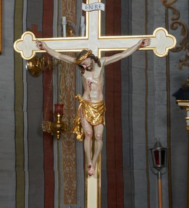Zdjęcie nr 1: Krzyż procesyjny na długim drzewcu o trójlistnych zakończeniach i złoconym obramowaniu. Figura Chrystusa umarłego ułożona w lekkim zwisie, przybita do krzyża trzema gwoździami, ze stopami założonymi prawa na lewą, nogami delikatnie ugiętymi w kolanach i głową przechyloną na prawe ramię. Twarz szczupła, głęboko rzeźbiona, okolona zarostem, usta otwarte, oczy zamknięte; włosy długie, opadające na ramiona, na głowie korona cierniowa. Ciało szczupłe o delikatnie zaznaczonej muskulaturze i śladami męki. Perizonium złocone, zawiązane na guz na prawym biodrze z silnie drapowanym, zwisającym końcem wzdłuż prawego uda. Nad głową Chrystusa wisi tabliczka w formie białej banderoli w czerwonej, wąskiej ramce z napisem „I.N.R.I.”. Polichromia ciała naturalistyczna, perizonium i korona złocone.

