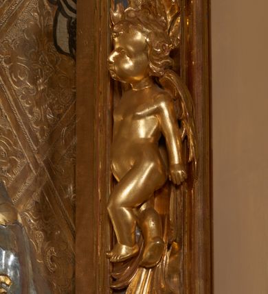 Zdjęcie nr 5: Obraz w kształcie stojącego prostokąta przedstawiający Matkę Boską z Dzieciątkiem. Maria została ukazana w półpostaci z Dzieciątkiem Jezus na prawym ręku, z gruszką w lewej dłoni, z głową delikatnie przechyloną na prawe ramię i wzrokiem skierowanym na widza. Dzieciątko ukazane jest w pozycji siedzącej, zwrócone trzy czwarte w lewo, lewą rączką sięga po gruszkę trzymaną przez Marię w dłoniach, prawą czyni gest błogosławieństwa. Na postacie Marii i Dzieciątka nałożone są metalowe, złocone i srebrzone sukienki, dekorowane w motywy floralne, a także korony w typie zamkniętym. Karnacje bardzo ciemne. Całość ukazana jest na złotym tle, w którym wyróżniają się koliste, złocone nimby wokół głów Marii i Dzieciątka oraz motywy roślinno-geometryczne zamknięte w rombach ukośnej kratownicy. Symetrycznie po bokach głowy Marii zostali przedstawieni konturowaniem dwaj aniołowie koronujący. Obraz umieszczony jest w profilowanej, złoconej i srebrzonej ramie, zdobionej rzeźbiarską, floralną dekoracją oraz parą aniołków na bokach ramy, ukazanych w postawie stojącej, zwróconych ku środkowi obrazu. 