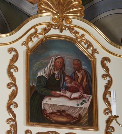 Zdjęcie nr 2: Obraz w kształcie stojącego prostokąta zamkniętego łukiem delikatnie wklęsło-wypukłym przedstawiający scenę Nauczania Marii. W centrum kompozycji ukazana Święta Rodzina: św. Anna, św. Joachim oraz mała Matka Boska zasiadający przy stole. Anna ubrana w zieloną suknię i biały welon założony na głowę, nad jej głową biały, okrągły nimb z gwiazdą w polu. Prawą ręką przytrzymuje księgę, a lewą obejmuje Marię. Joachim ukazany za Matką Boską, ubrany w białą koszulę, w brązową suknię i niebieski płaszcz. Maria w różowej szacie podtrzymuje dłońmi otwartą na stole księgę, wokół jej głowy delikatny promienisty, biały nimb. Stół nakryty białym obrusem, na którym leżą: po lewej stronie jabłko, po prawej kiście winnego grona i gałązki róż. Przy dolnej krawędzi złoty rozciągnięty na boki kartusz  z napisem „Anna Joahim i Marya”. Szeroka gama barwna, z przewagą zimnych kolorów. W tle daleki krajobraz, z wysokim drzewem, niebo jasnobłękitne.