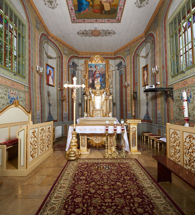 Zdjęcie nr 5: Ława ustawiona na niskim prostopadłościennym podeście, jednorzędowa, trójosobowa, z zapleckiem i z przedpiersiem, zwieńczona dwiema wolutami spinającymi pośrodku palmetę oraz małymi wazonikami ustawionymi na skrajach zaplecka. Przedpiersie przedzielone na trzy płyciny w kształcie stojących prostokątów zdobione w polu stylizowaną wicią z motywami ornamentu pseudoregencyjnego. Zaplecek przedzielony na dwie strefy; w dolnej zdobiony trzema kwadratowymi płycinami, w górnej płyciną w kształcie leżącego prostokąta zamkniętego od góry łukiem wklęsło-wypukłym. 
