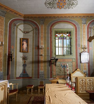 Zdjęcie nr 7: Ława ustawiona na niskim prostopadłościennym podeście, jednorzędowa, trójosobowa, z zapleckiem i z przedpiersiem, zwieńczona dwiema wolutami spinającymi pośrodku palmetę oraz małymi wazonikami ustawionymi na skrajach zaplecka. Przedpiersie przedzielone na trzy płyciny w kształcie stojących prostokątów zdobione w polu stylizowaną wicią z motywami ornamentu pseudoregencyjnego. Zaplecek przedzielony na dwie strefy; w dolnej zdobiony trzema kwadratowymi płycinami, w górnej płyciną w kształcie leżącego prostokąta zamkniętego od góry łukiem wklęsło-wypukłym. 
