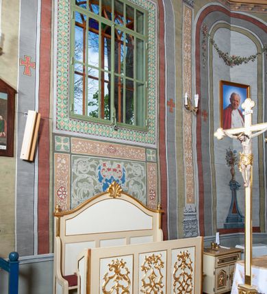 Zdjęcie nr 2: Ława ustawiona na niskim prostopadłościennym podeście, jednorzędowa, trójosobowa, z zapleckiem i z przedpiersiem, zwieńczona dwiema wolutami spinającymi pośrodku palmetę oraz małymi wazonikami ustawionymi na skrajach zaplecka. Przedpiersie przedzielone na trzy płyciny w kształcie stojących prostokątów zdobione w polu stylizowaną wicią z motywami ornamentu pseudoregencyjnego. Zaplecek przedzielony na dwie strefy; w dolnej zdobiony trzema kwadratowymi płycinami, w górnej płyciną w kształcie leżącego prostokąta zamkniętego od góry łukiem wklęsło-wypukłym. 
