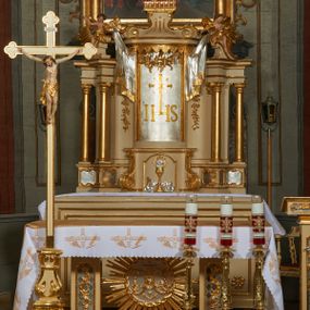 Zdjęcie nr 1: Ołtarz murowany, w formie prostopadłościanu. Mensa drewniana z portatylem. Antependium złocone, w formie leżącego prostokąta w profilowanej ramie; w polu środkowym znajduje się monogram IHS, a w narożach rozety. Tabernakulum z wypukłą ścianką frontową, po bokach ujęte jest spływami wolutowymi na cokołach, dekorowanymi ornamentem rocaille'owym. Drzwiczki w formie stojącego prostokąta zamkniętego łukiem nadwieszonym; w polu mają płaskorzeźbiony kielich wśród obłoków, z hostią w glorii. Tron wystawienia architektoniczny, trójosiowy, jednokondygnacyjny ze zwieńczeniem. Oś środkowa wypukła, wyższa niż boczne, zamknięta jest profilowanym gzymsem. Tron ujmują dwie pary kolumn na ukośnie ustawionych cokołach. Podpory dźwigają pełne belkowanie wyłamane na ich osi. Na linii wewnętrznych kolumn znajduje się przerywany przyczółek z siedzącymi aniołkami podtrzymującymi srebrzony paludament. Zwieńczenie ma formę cokołu, ujętego spływami wolutowymi; na którym ustawiona jest księga zamknięta na siedem pięczęci, na niej postawiona srebrzona waza z kwiatami oraz biały baranek z chorągwią paschalną. Na cokołach wspierających kolumny znajdują się rocaille'owe płyciny z lusterkami. Oś środkowa flankowana jest kwiatowymi zwisami. Tron wystawienia posiada obrotową wnękę z gzymsem wieńczącym, ozdobionym lambrekinem: awers wklęsły, posrebrzany, z rytym monogramem IHS oraz krzyżem i wiązkami promieni, powyżej rzeźbiony rocaille; rewers w kształcie niszy, dekorowany rytą, złoconą kratką i rzeźbionym, srebrzonym rocaille'm. Struktura polichromowana na beżowo, kolumny, ornamentyka i profilowania złocone, detale srebrzone. Za ołtarzem, na wschodniej ścianie prezbiterium wisi obraz św. Erazma w złoconej, profilowanej ramie, w formie stojącego prostokąta, zamkniętego nadwieszonym łukiem półkolistym. Rama dekorowana jest w narożach i na bokach stylizowanymi palmetami. Nad obrazem, w podłuczu znajduje się płaskorzeźbiona korona otwarta z dwoma piórami.