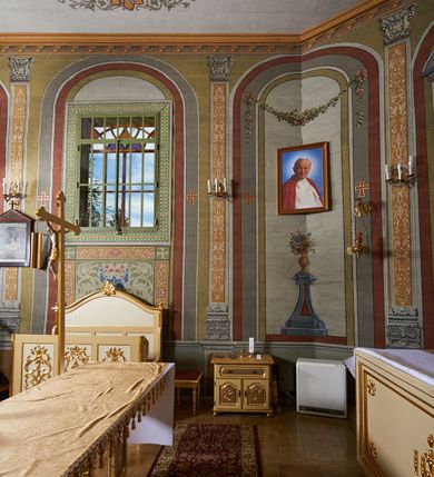 Zdjęcie nr 12: Polichromia umieszczona na ścianach prezbiterium naśladuje architekturę. Składa się z czterech prostokątnych płaszczyzn, zamkniętych od góry półkoliście, ujętych w pasach w kolorze oliwkowozielonym i czerwonym, oddzielonych od siebie pilastrami. Trzony pilastrów dekorowane są ornamentem ze stylizowanych liści i kwiatów; bazy i kapitele są z motywami zoomorficznymi i roślinnymi w kolorze szarym. W dolnych częściach płaszczyzn, przy ołtarzu namalowano kwiatony, w górnych częściach kwieciste girlandy. Na ścianie wschodniej znajduje się namalowana supraporta z częściowo zachowanym napisem „AEDIFICATUM ET DEDIC(TUM)?”. Wokół obrazu św. Erazma zawieszonego na ścianie wschodniej namalowano pseudorokokową ramę w kolorze złotym. Dołem biegnie pas lamperii w kolorze białym. Na stropie, w centralnej części kompozycji ukazana jest scena Zwiastowania, namalowana w technice olejnej na płótnie. Po lewej stronie znajduje się Matka Boska w różowej sukni i niebieskim płaszczu, a po prawej św. Archanioł Gabriel w białej sukni i ugrowym płaszczu. Nad nimi w promienistej glorii została ukazana gołębica Ducha Świętego. Scena namalowana jest w architektonicznym wnętrzu. Tło jasnoszare. Obraz ujmuje bordiura z powtarzających się elementów geometryczno-roślinnych. Analogicznie jak w prezbiterium w nawie głównej znajduje się pięć architektonicznych płaszczyzn. Polichromie umieszczone na ścianach południowej i północnej stanowią swoje lustrzane odbicie. W górnej części płaszczyzn namalowano girlandy oraz dwie tarcze herbowe w supraportach. Dodatkowo w płaszczyznach podokiennych namalowano wijącą się dywanową kompozycję ze stylizowanych liści i kwiatów. Pod chórem umieszczone są dwa zacheusze w pseudorokokowej ramie. U dołu nawę obiega pas lamperii w kolorze zielonym. Na stropie, w części centralnej znajduje się obraz przedstawiający Przemienienie Pańskie. Tło jasnoszare. Obraz ujmuje bordiura z powtarzających się elementów geometryczno-roślinnych.