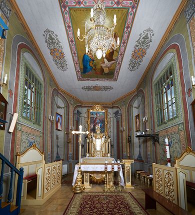 Zdjęcie nr 11: Polichromia umieszczona na ścianach prezbiterium naśladuje architekturę. Składa się z czterech prostokątnych płaszczyzn, zamkniętych od góry półkoliście, ujętych w pasach w kolorze oliwkowozielonym i czerwonym, oddzielonych od siebie pilastrami. Trzony pilastrów dekorowane są ornamentem ze stylizowanych liści i kwiatów; bazy i kapitele są z motywami zoomorficznymi i roślinnymi w kolorze szarym. W dolnych częściach płaszczyzn, przy ołtarzu namalowano kwiatony, w górnych częściach kwieciste girlandy. Na ścianie wschodniej znajduje się namalowana supraporta z częściowo zachowanym napisem „AEDIFICATUM ET DEDIC(TUM)?”. Wokół obrazu św. Erazma zawieszonego na ścianie wschodniej namalowano pseudorokokową ramę w kolorze złotym. Dołem biegnie pas lamperii w kolorze białym. Na stropie, w centralnej części kompozycji ukazana jest scena Zwiastowania, namalowana w technice olejnej na płótnie. Po lewej stronie znajduje się Matka Boska w różowej sukni i niebieskim płaszczu, a po prawej św. Archanioł Gabriel w białej sukni i ugrowym płaszczu. Nad nimi w promienistej glorii została ukazana gołębica Ducha Świętego. Scena namalowana jest w architektonicznym wnętrzu. Tło jasnoszare. Obraz ujmuje bordiura z powtarzających się elementów geometryczno-roślinnych. Analogicznie jak w prezbiterium w nawie głównej znajduje się pięć architektonicznych płaszczyzn. Polichromie umieszczone na ścianach południowej i północnej stanowią swoje lustrzane odbicie. W górnej części płaszczyzn namalowano girlandy oraz dwie tarcze herbowe w supraportach. Dodatkowo w płaszczyznach podokiennych namalowano wijącą się dywanową kompozycję ze stylizowanych liści i kwiatów. Pod chórem umieszczone są dwa zacheusze w pseudorokokowej ramie. U dołu nawę obiega pas lamperii w kolorze zielonym. Na stropie, w części centralnej znajduje się obraz przedstawiający Przemienienie Pańskie. Tło jasnoszare. Obraz ujmuje bordiura z powtarzających się elementów geometryczno-roślinnych.