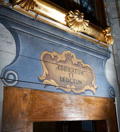 Zdjęcie nr 8: Polichromia umieszczona na ścianach prezbiterium naśladuje architekturę. Składa się z czterech prostokątnych płaszczyzn, zamkniętych od góry półkoliście, ujętych w pasach w kolorze oliwkowozielonym i czerwonym, oddzielonych od siebie pilastrami. Trzony pilastrów dekorowane są ornamentem ze stylizowanych liści i kwiatów; bazy i kapitele są z motywami zoomorficznymi i roślinnymi w kolorze szarym. W dolnych częściach płaszczyzn, przy ołtarzu namalowano kwiatony, w górnych częściach kwieciste girlandy. Na ścianie wschodniej znajduje się namalowana supraporta z częściowo zachowanym napisem „AEDIFICATUM ET DEDIC(TUM)?”. Wokół obrazu św. Erazma zawieszonego na ścianie wschodniej namalowano pseudorokokową ramę w kolorze złotym. Dołem biegnie pas lamperii w kolorze białym. Na stropie, w centralnej części kompozycji ukazana jest scena Zwiastowania, namalowana w technice olejnej na płótnie. Po lewej stronie znajduje się Matka Boska w różowej sukni i niebieskim płaszczu, a po prawej św. Archanioł Gabriel w białej sukni i ugrowym płaszczu. Nad nimi w promienistej glorii została ukazana gołębica Ducha Świętego. Scena namalowana jest w architektonicznym wnętrzu. Tło jasnoszare. Obraz ujmuje bordiura z powtarzających się elementów geometryczno-roślinnych. Analogicznie jak w prezbiterium w nawie głównej znajduje się pięć architektonicznych płaszczyzn. Polichromie umieszczone na ścianach południowej i północnej stanowią swoje lustrzane odbicie. W górnej części płaszczyzn namalowano girlandy oraz dwie tarcze herbowe w supraportach. Dodatkowo w płaszczyznach podokiennych namalowano wijącą się dywanową kompozycję ze stylizowanych liści i kwiatów. Pod chórem umieszczone są dwa zacheusze w pseudorokokowej ramie. U dołu nawę obiega pas lamperii w kolorze zielonym. Na stropie, w części centralnej znajduje się obraz przedstawiający Przemienienie Pańskie. Tło jasnoszare. Obraz ujmuje bordiura z powtarzających się elementów geometryczno-roślinnych.