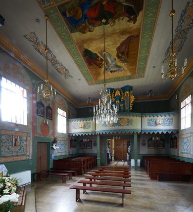 Zdjęcie nr 5: Polichromia umieszczona na ścianach prezbiterium naśladuje architekturę. Składa się z czterech prostokątnych płaszczyzn, zamkniętych od góry półkoliście, ujętych w pasach w kolorze oliwkowozielonym i czerwonym, oddzielonych od siebie pilastrami. Trzony pilastrów dekorowane są ornamentem ze stylizowanych liści i kwiatów; bazy i kapitele są z motywami zoomorficznymi i roślinnymi w kolorze szarym. W dolnych częściach płaszczyzn, przy ołtarzu namalowano kwiatony, w górnych częściach kwieciste girlandy. Na ścianie wschodniej znajduje się namalowana supraporta z częściowo zachowanym napisem „AEDIFICATUM ET DEDIC(TUM)?”. Wokół obrazu św. Erazma zawieszonego na ścianie wschodniej namalowano pseudorokokową ramę w kolorze złotym. Dołem biegnie pas lamperii w kolorze białym. Na stropie, w centralnej części kompozycji ukazana jest scena Zwiastowania, namalowana w technice olejnej na płótnie. Po lewej stronie znajduje się Matka Boska w różowej sukni i niebieskim płaszczu, a po prawej św. Archanioł Gabriel w białej sukni i ugrowym płaszczu. Nad nimi w promienistej glorii została ukazana gołębica Ducha Świętego. Scena namalowana jest w architektonicznym wnętrzu. Tło jasnoszare. Obraz ujmuje bordiura z powtarzających się elementów geometryczno-roślinnych. Analogicznie jak w prezbiterium w nawie głównej znajduje się pięć architektonicznych płaszczyzn. Polichromie umieszczone na ścianach południowej i północnej stanowią swoje lustrzane odbicie. W górnej części płaszczyzn namalowano girlandy oraz dwie tarcze herbowe w supraportach. Dodatkowo w płaszczyznach podokiennych namalowano wijącą się dywanową kompozycję ze stylizowanych liści i kwiatów. Pod chórem umieszczone są dwa zacheusze w pseudorokokowej ramie. U dołu nawę obiega pas lamperii w kolorze zielonym. Na stropie, w części centralnej znajduje się obraz przedstawiający Przemienienie Pańskie. Tło jasnoszare. Obraz ujmuje bordiura z powtarzających się elementów geometryczno-roślinnych.