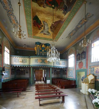 Zdjęcie nr 4: Polichromia umieszczona na ścianach prezbiterium naśladuje architekturę. Składa się z czterech prostokątnych płaszczyzn, zamkniętych od góry półkoliście, ujętych w pasach w kolorze oliwkowozielonym i czerwonym, oddzielonych od siebie pilastrami. Trzony pilastrów dekorowane są ornamentem ze stylizowanych liści i kwiatów; bazy i kapitele są z motywami zoomorficznymi i roślinnymi w kolorze szarym. W dolnych częściach płaszczyzn, przy ołtarzu namalowano kwiatony, w górnych częściach kwieciste girlandy. Na ścianie wschodniej znajduje się namalowana supraporta z częściowo zachowanym napisem „AEDIFICATUM ET DEDIC(TUM)?”. Wokół obrazu św. Erazma zawieszonego na ścianie wschodniej namalowano pseudorokokową ramę w kolorze złotym. Dołem biegnie pas lamperii w kolorze białym. Na stropie, w centralnej części kompozycji ukazana jest scena Zwiastowania, namalowana w technice olejnej na płótnie. Po lewej stronie znajduje się Matka Boska w różowej sukni i niebieskim płaszczu, a po prawej św. Archanioł Gabriel w białej sukni i ugrowym płaszczu. Nad nimi w promienistej glorii została ukazana gołębica Ducha Świętego. Scena namalowana jest w architektonicznym wnętrzu. Tło jasnoszare. Obraz ujmuje bordiura z powtarzających się elementów geometryczno-roślinnych. Analogicznie jak w prezbiterium w nawie głównej znajduje się pięć architektonicznych płaszczyzn. Polichromie umieszczone na ścianach południowej i północnej stanowią swoje lustrzane odbicie. W górnej części płaszczyzn namalowano girlandy oraz dwie tarcze herbowe w supraportach. Dodatkowo w płaszczyznach podokiennych namalowano wijącą się dywanową kompozycję ze stylizowanych liści i kwiatów. Pod chórem umieszczone są dwa zacheusze w pseudorokokowej ramie. U dołu nawę obiega pas lamperii w kolorze zielonym. Na stropie, w części centralnej znajduje się obraz przedstawiający Przemienienie Pańskie. Tło jasnoszare. Obraz ujmuje bordiura z powtarzających się elementów geometryczno-roślinnych.