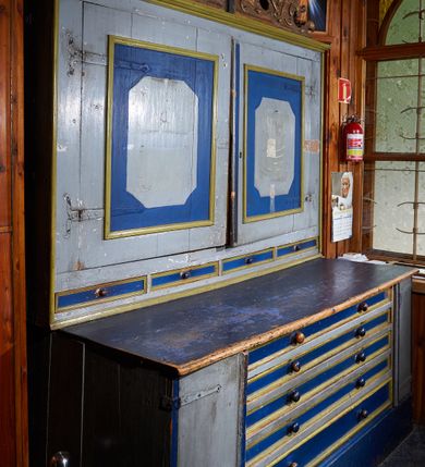 Zdjęcie nr 1: Komoda prostopadłościenna, ustawiona na wysokim cokole, po bokach z dwiema jednodrzwiowymi szafkami, pośrodku pięć niskich szuflad na ornaty. Nakryta blatem o profilowanej od przodu krawędzi. Drzwiczki osadzone są na metalowych, kutych zawiasach. Szuflady posiadają lica z profilowanymi listwami i parami kulistych, toczonych uchwytów. Drzwiczki zewnętrznych szafek posiadają profilowane listwy przymykowe. Nadstawka dwudrzwiowa, w dolnej części z czterema, niskimi szufladami, obitymi profilowanymi listwami, z toczonymi, kulistymi uchwytami; zwieńczona profilowanym gzymsem. Drzwi osadzone na metalowych, kutych zawiasach pasowych, w polu zdobione malowanymi płycinami w kształcie stojącego prostokąta o ściętych narożach. Zwieńczenie w formie rzeźbionej wici roślinnej miejscami wywiniętej kartuszowo. Pośrodku szczytu osadzony jest krucyfiks z figurą Chrystusa w typie Cristo morte. Szafa malowana olejno w kolorze jasnoniebieskim, detal na niebiesko, listwy na beżowo.