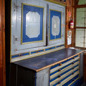 Zdjęcie nr 1: Komoda prostopadłościenna, ustawiona na wysokim cokole, po bokach z dwiema jednodrzwiowymi szafkami, pośrodku pięć niskich szuflad na ornaty. Nakryta blatem o profilowanej od przodu krawędzi. Drzwiczki osadzone są na metalowych, kutych zawiasach. Szuflady posiadają lica z profilowanymi listwami i parami kulistych, toczonych uchwytów. Drzwiczki zewnętrznych szafek posiadają profilowane listwy przymykowe. Nadstawka dwudrzwiowa, w dolnej części z czterema, niskimi szufladami, obitymi profilowanymi listwami, z toczonymi, kulistymi uchwytami; zwieńczona profilowanym gzymsem. Drzwi osadzone na metalowych, kutych zawiasach pasowych, w polu zdobione malowanymi płycinami w kształcie stojącego prostokąta o ściętych narożach. Zwieńczenie w formie rzeźbionej wici roślinnej miejscami wywiniętej kartuszowo. Pośrodku szczytu osadzony jest krucyfiks z figurą Chrystusa w typie Cristo morte. Szafa malowana olejno w kolorze jasnoniebieskim, detal na niebiesko, listwy na beżowo.