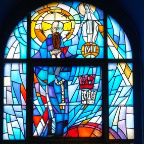 Zdjęcie nr 1: Witraż o wykroju prostokąta stojącego zakończonego łukiem półkolistym. Kompozycja dwustrefowa, w dolnej nierozpoznany święty zwrócony trzy czwarte w lewo z krzyżem w dłoniach na wysokości piersi. Twarz szeroka, ukazana z prawego profilu o wyrazistych rysach z dużymi oczami i długim nosem. Zakonnik ubrany jest w habit. W górnej strefie św. Maksymilian Maria Kolbe przed Matką Boską. Święty ukazany jest frontalnie z dłońmi złożonymi w geście modlitwy. Twarz szeroka, szczupła z wyraźnie podkreślonymi rysami twarzy, okolona długą, brązową brodą. Na nosie ma założone proste, okrągłe okulary; włosy krótkie. Wokół głowy ma okrągły nimb otoczony drutem kolczastym, na lewym ramieniu trójkątną naszywkę z napisem „P”. Po prawej stronie witrażu ukazane są dwie korony: biała i czerwona, a powyżej Matka Boska na złotym globie z literami „MI”. Maria ukazana sumarycznie z rękami rozłożonymi szeroko na boki, delikatnie pochylona do przodu. Ubrana jest w długą suknię oraz płaszcz założony na głowę i spływający wzdłuż boków. Witraż utrzymany w chłodnej, niebieskiej kolorystyce, ożywiony kolorami czerwieni i złota wokół głów postaci. 

