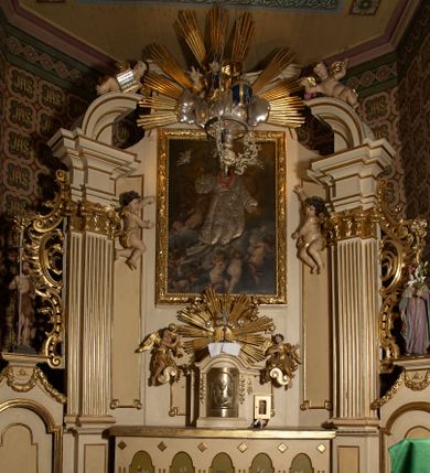 Zdjęcie nr 1: Ołtarz typu architektonicznego, jednokondygnacyjny, jednoosiowy, z bramkami i ze zwieńczeniem w formie glorii. Mensa prostopadłościenna, na ściance przedniej zdobiona rzędem płycin w kształcie stojącego prostokąta, zamkniętych łukiem nadwieszonym. W polu środkowej płyciny malowany krzyż grecki, w pozostałych gałązki białych lilii. Tabernakulum w formie prostopadłościennej skrzynki, z częścią środkową wysuniętą do przodu, zamknięte od góry profilowanym gzymsem w kształcie łuku nadwieszonego. Drzwiczki metalowe, pośrodku dekorowane płyciną w kształcie stojącego prostokąta, zamkniętego łukiem nadwieszonym, w polu kielich z hostią, kłosy zboża i kiście winogron. Nad tabernakulum tron wystawienia na tle promienistej glorii z monogramem IHS pośrodku, po bokach flankowany przez parę aniołków usadzonych na wolutach. Retabulum ustawione na wysokim cokole, ujęte parą zdwojonych, ukośnie ustawionych pilastrów podtrzymujących przerywane belkowanie, na którym usadzona para putt. Trzony pilastrów zdobione kanelowaniem, kapitele stylizowane, kompozytowe. Putto po lewej stronie z otwartą księgą, w której znajduje się napis: „Assu/pta / est / Maria / in / Caelum”. W polu głównym nastawy, na wysokości dwóch trzecich obraz z przedstawieniem Wniebowzięcia Najświętszej Marii Panny. Po bokach obrazu para rzeźbionych aniołków. W zwieńczeniu płaskorzeźba Boga Ojca na tle promienistej glorii. Drzwiczki bramek zdobione płyciną w kształcie stojącego prostokąta, zamkniętą od góry łukiem nadwieszonym. Zwieńczenie bramek zdobione festonem z liści wawrzynu, zakończone niskim cokołem, na którym ustawione rzeźby: po lewej św. Jana Chrzciciela, po prawej św. Kazimierza. Uszy ażurowe w postaci ornamentu rocaille'owego. Struktura ołtarza w kolorze beżowym, ornamentyka, detale i profilowania złocone.