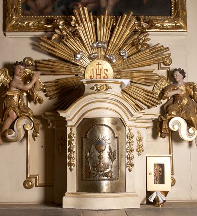 Zdjęcie nr 11: Ołtarz typu architektonicznego, jednokondygnacyjny, jednoosiowy, z bramkami i ze zwieńczeniem w formie glorii. Mensa prostopadłościenna, na ściance przedniej zdobiona rzędem płycin w kształcie stojącego prostokąta, zamkniętych łukiem nadwieszonym. W polu środkowej płyciny malowany krzyż grecki, w pozostałych gałązki białych lilii. Tabernakulum w formie prostopadłościennej skrzynki, z częścią środkową wysuniętą do przodu, zamknięte od góry profilowanym gzymsem w kształcie łuku nadwieszonego. Drzwiczki metalowe, pośrodku dekorowane płyciną w kształcie stojącego prostokąta, zamkniętego łukiem nadwieszonym, w polu kielich z hostią, kłosy zboża i kiście winogron. Nad tabernakulum tron wystawienia na tle promienistej glorii z monogramem IHS pośrodku, po bokach flankowany przez parę aniołków usadzonych na wolutach. Retabulum ustawione na wysokim cokole, ujęte parą zdwojonych, ukośnie ustawionych pilastrów podtrzymujących przerywane belkowanie, na którym usadzona para putt. Trzony pilastrów zdobione kanelowaniem, kapitele stylizowane, kompozytowe. Putto po lewej stronie z otwartą księgą, w której znajduje się napis: „Assu/pta / est / Maria / in / Caelum”. W polu głównym nastawy, na wysokości dwóch trzecich obraz z przedstawieniem Wniebowzięcia Najświętszej Marii Panny. Po bokach obrazu para rzeźbionych aniołków. W zwieńczeniu płaskorzeźba Boga Ojca na tle promienistej glorii. Drzwiczki bramek zdobione płyciną w kształcie stojącego prostokąta, zamkniętą od góry łukiem nadwieszonym. Zwieńczenie bramek zdobione festonem z liści wawrzynu, zakończone niskim cokołem, na którym ustawione rzeźby: po lewej św. Jana Chrzciciela, po prawej św. Kazimierza. Uszy ażurowe w postaci ornamentu rocaille&#039;owego. Struktura ołtarza w kolorze beżowym, ornamentyka, detale i profilowania złocone.