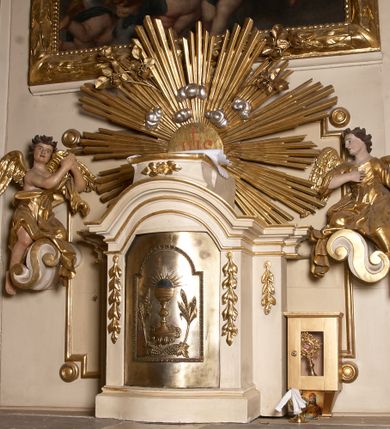 Zdjęcie nr 12: Ołtarz typu architektonicznego, jednokondygnacyjny, jednoosiowy, z bramkami i ze zwieńczeniem w formie glorii. Mensa prostopadłościenna, na ściance przedniej zdobiona rzędem płycin w kształcie stojącego prostokąta, zamkniętych łukiem nadwieszonym. W polu środkowej płyciny malowany krzyż grecki, w pozostałych gałązki białych lilii. Tabernakulum w formie prostopadłościennej skrzynki, z częścią środkową wysuniętą do przodu, zamknięte od góry profilowanym gzymsem w kształcie łuku nadwieszonego. Drzwiczki metalowe, pośrodku dekorowane płyciną w kształcie stojącego prostokąta, zamkniętego łukiem nadwieszonym, w polu kielich z hostią, kłosy zboża i kiście winogron. Nad tabernakulum tron wystawienia na tle promienistej glorii z monogramem IHS pośrodku, po bokach flankowany przez parę aniołków usadzonych na wolutach. Retabulum ustawione na wysokim cokole, ujęte parą zdwojonych, ukośnie ustawionych pilastrów podtrzymujących przerywane belkowanie, na którym usadzona para putt. Trzony pilastrów zdobione kanelowaniem, kapitele stylizowane, kompozytowe. Putto po lewej stronie z otwartą księgą, w której znajduje się napis: „Assu/pta / est / Maria / in / Caelum”. W polu głównym nastawy, na wysokości dwóch trzecich obraz z przedstawieniem Wniebowzięcia Najświętszej Marii Panny. Po bokach obrazu para rzeźbionych aniołków. W zwieńczeniu płaskorzeźba Boga Ojca na tle promienistej glorii. Drzwiczki bramek zdobione płyciną w kształcie stojącego prostokąta, zamkniętą od góry łukiem nadwieszonym. Zwieńczenie bramek zdobione festonem z liści wawrzynu, zakończone niskim cokołem, na którym ustawione rzeźby: po lewej św. Jana Chrzciciela, po prawej św. Kazimierza. Uszy ażurowe w postaci ornamentu rocaille'owego. Struktura ołtarza w kolorze beżowym, ornamentyka, detale i profilowania złocone.