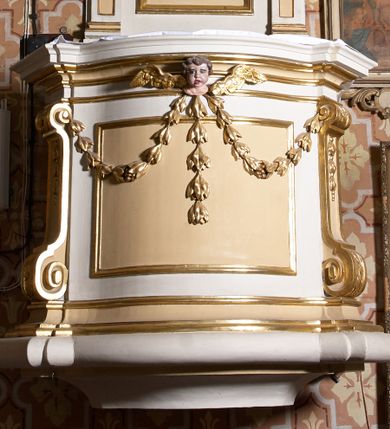 Zdjęcie nr 1: Ambona przyścienna, wisząca. Kosz czworoboczny, wybrzuszony z przodu na rzucie łuku odcinkowego. Na narożach flankowany pilastrami, zdobionymi wolutami. Ścianki korpusu dekorowane płycinami w kształcie leżącego prostokąta. Parapet w formie profilowanego gzymsu. Na ściance przedniej, w partii gzymsu, uskrzydlona główka aniołka, spod której zwisa girlanda utworzona z kampanul. Kosz ambony ścięty od dołu prosto. Zaplecek ujęty parą pilastrów, z płycinami zdobionymi w części górnej zwisami ze stylizowanych kampanul, z nałożonymi u szczytu konsolkami, pełniącymi funkcję kapiteli, które wspierają odcinki przerwanego belkowania. Pośrodku zaplecka płycina w kształcie stojącego prostokąta zamknięta od góry łukiem wklęsłym, nad nią stylizowana muszla palmetowa, poniżej zwis utworzony z gałązki róży. Zwieńczenie w formie promienistej glorii, usytuowanej pomiędzy dwiema wolutami osadzonymi na odcinkach belkowania na osiach pilastrów. Gloria utworzona z rozszerzających się wiązek promieni o nierównej długości, pośrodku gołębica Ducha Świętego w otoku chmur. Struktura ambony malowana w kolorze białym, płyciny i detale w kolorze beżowo-kremowym, ornamentyka i profilowania złocone i srebrzone.
